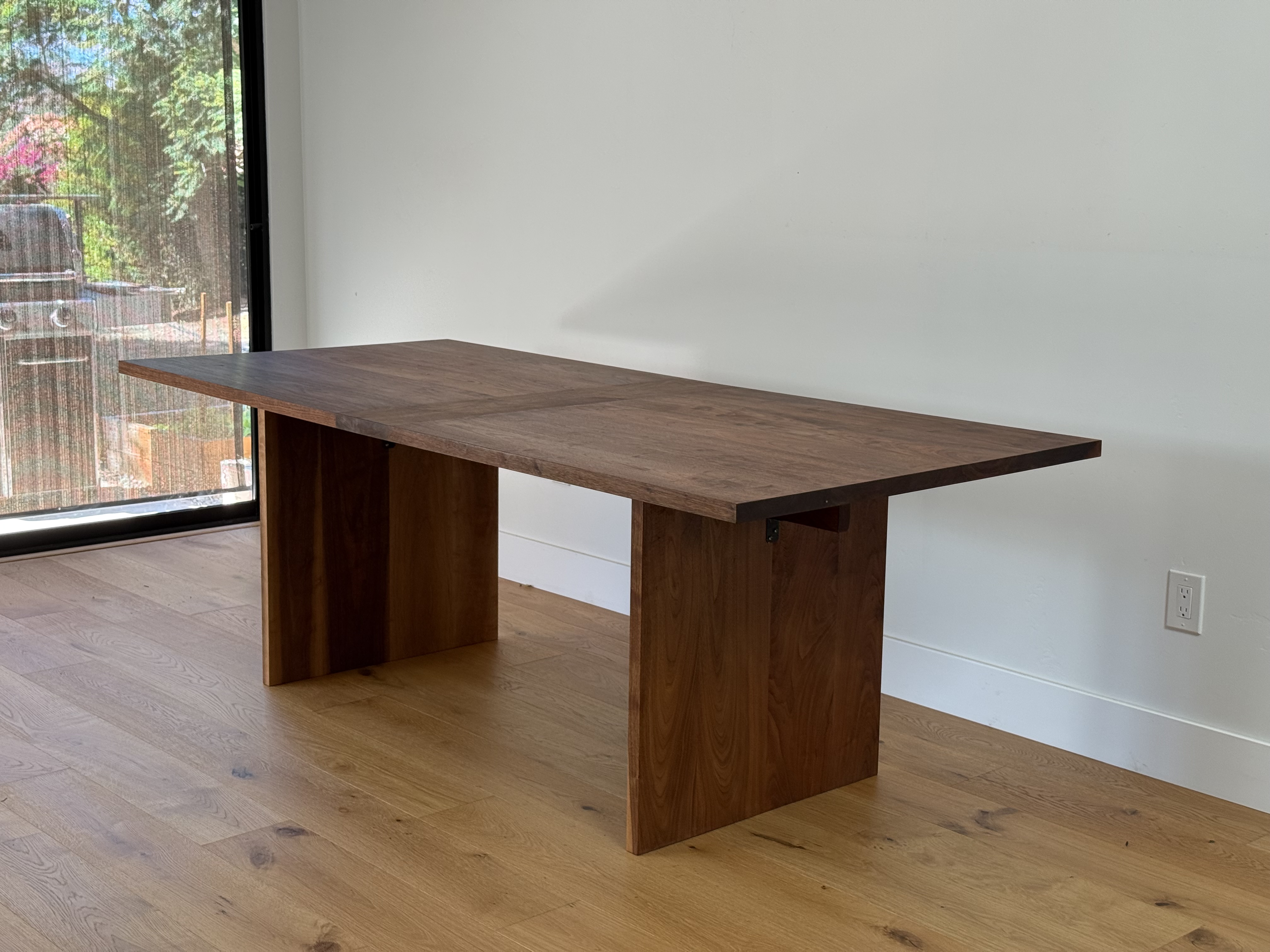 Walnut dining table with plank top and paneled trestle legs, in a room with natural light
